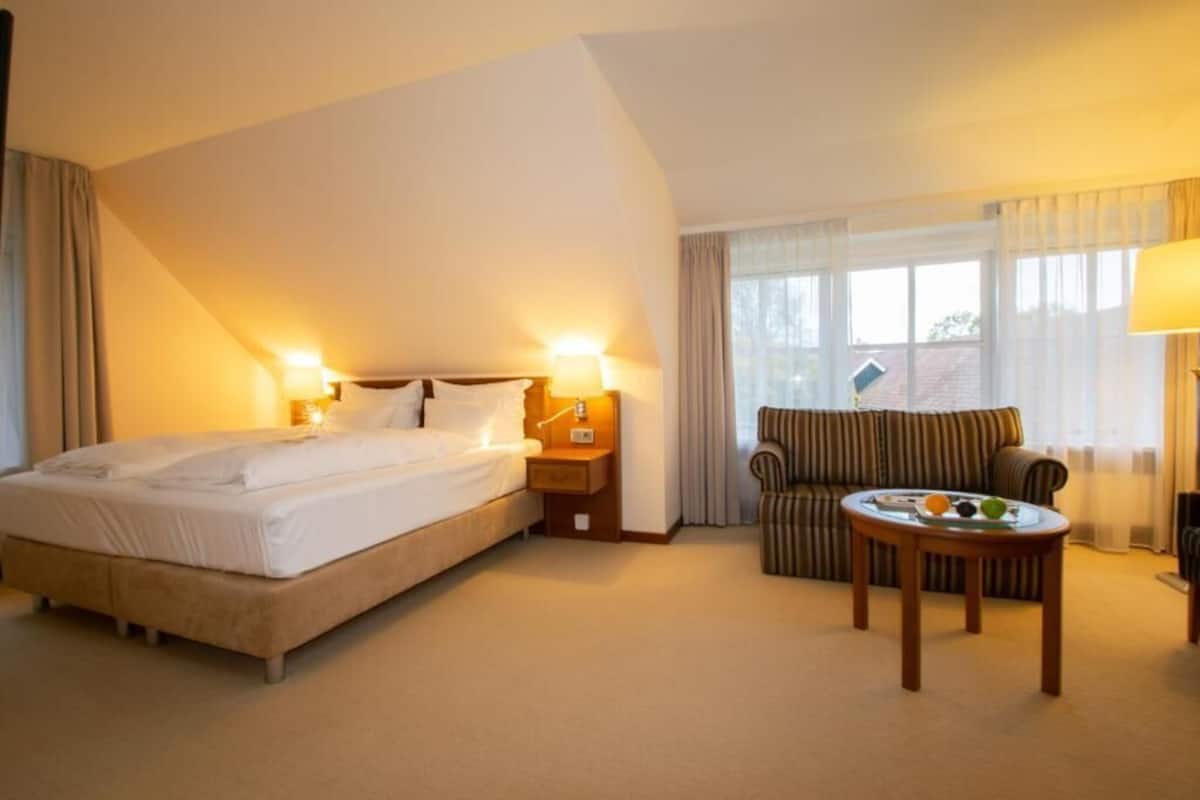 Une chambre d’hôtel avec un lit, des lampes de chevet, un canapé, une petite table avec des fruits et une fenêtre avec des rideaux.