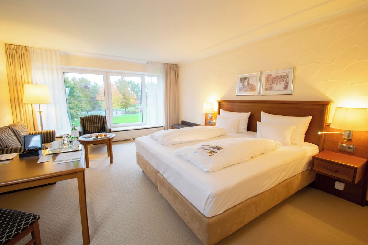 Une chambre d’hôtel avec un grand lit, un bureau avec un ordinateur portable, une chaise et une fenêtre donnant sur des arbres.