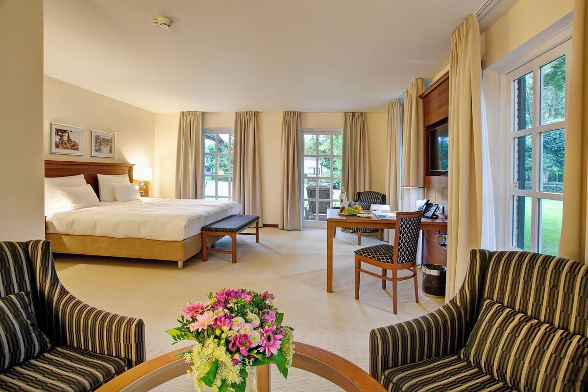 Une chambre d’hôtel avec un lit, un bureau, deux fauteuils, une table basse ornée de fleurs et de grandes fenêtres avec des rideaux.
