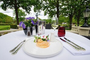A dining table set outdoors with a white tablecloth, a plate of food, silverware, a wine glass, and a bottle of wine. - Fletcher Parkhotel De Wiemsel (Ootmarsum)