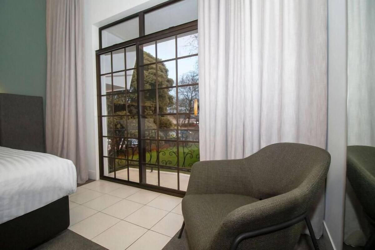 A hotel room with a bed, a chair, a large sliding glass door with a view of a garden, and a tiled floor.