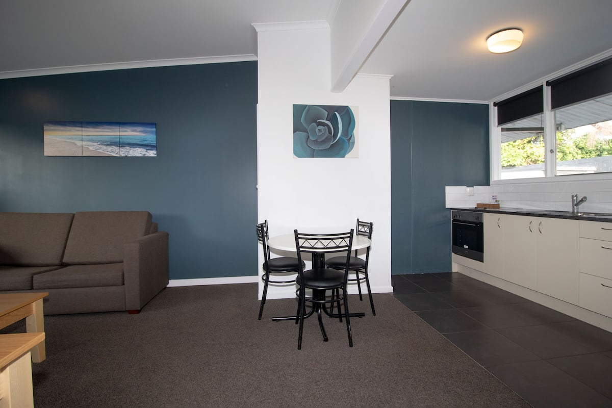 Een moderne woonkamer met een grijze bank, een eettafel met vier stoelen en een keuken met witte kasten en een raam.