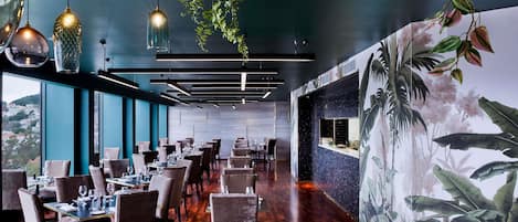 Interior de restaurante con suelo de madera, lámparas colgantes y un mural de follaje tropical en la pared.