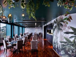 A restaurant interior with wooden flooring, hanging pendant lights, and a mural of tropical foliage on the wall.