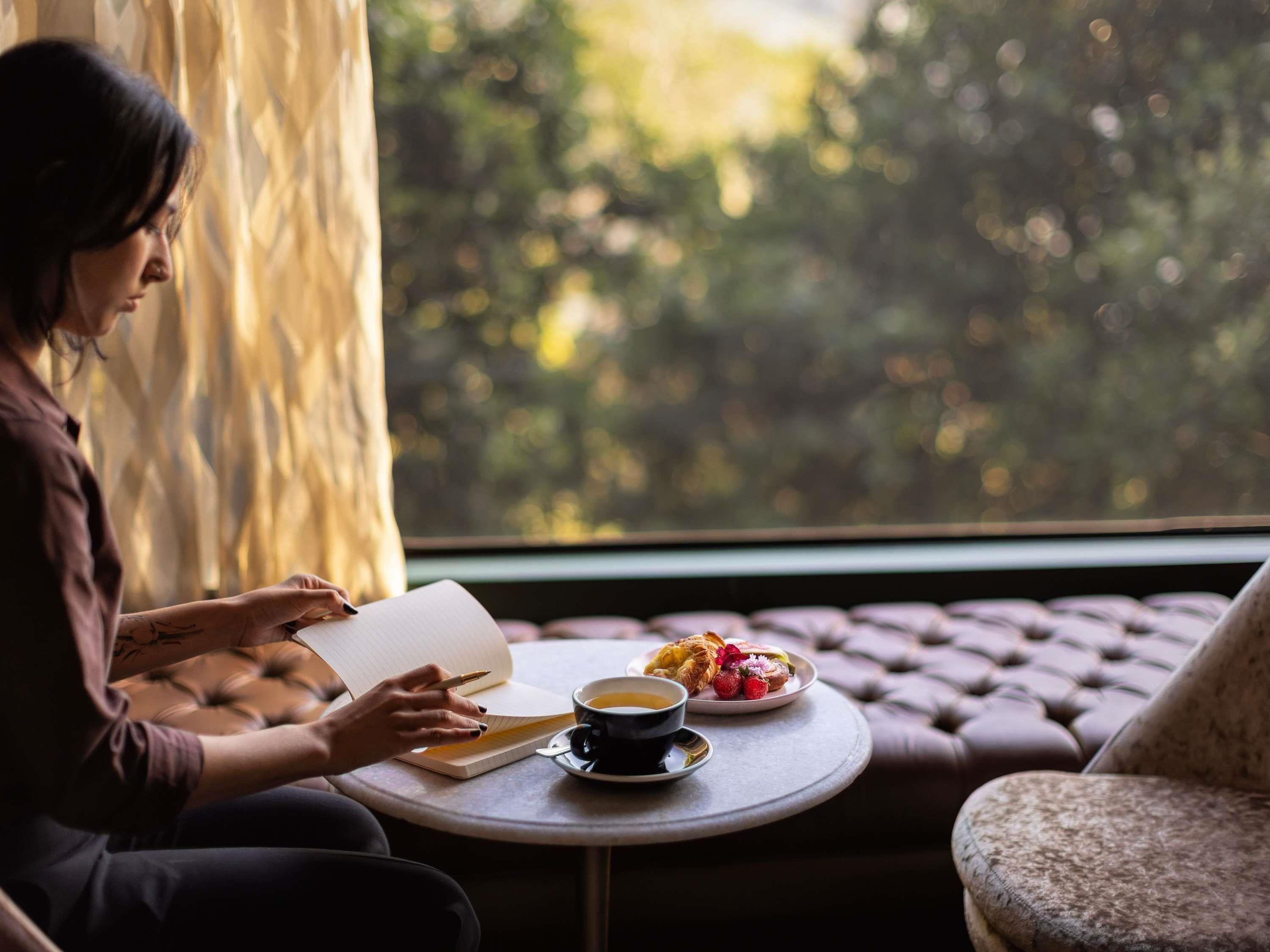 Une personne assise à une table ronde, devant un carnet, une tasse de café et une assiette de nourriture.