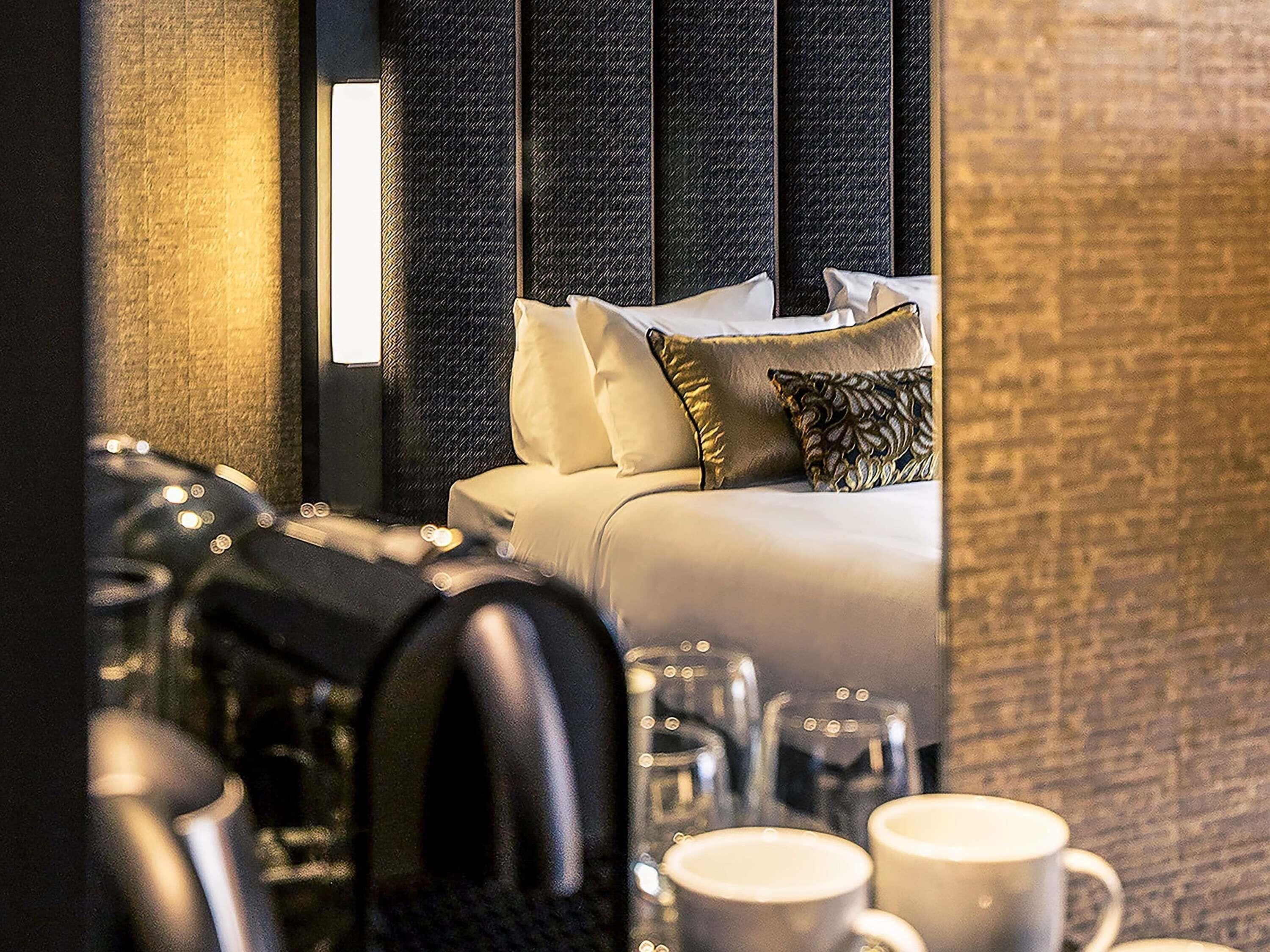 A modern hotel room with a bed, pillows, and a nightstand with glasses and a cup.