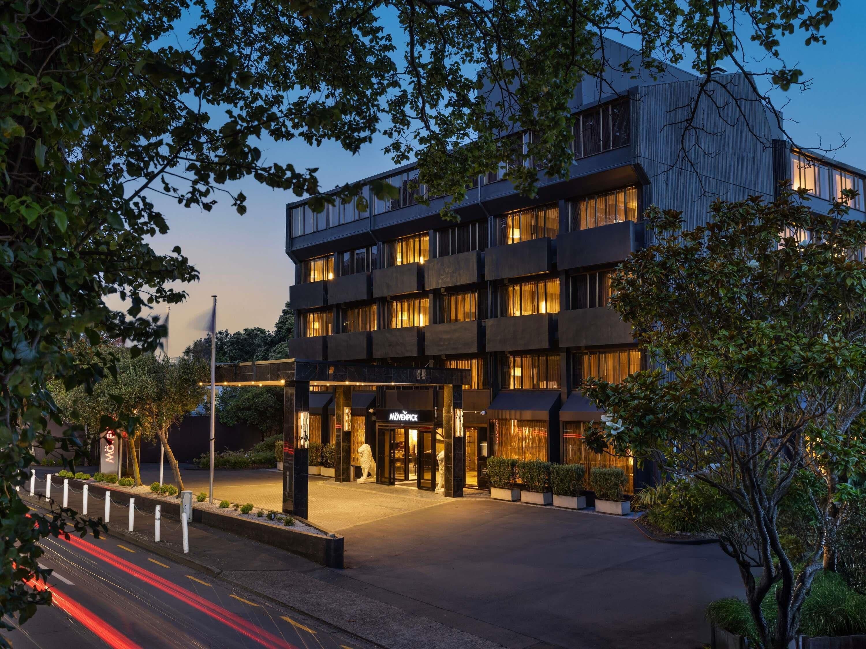Um hotel moderno com vários andares, entrada bem iluminada e área ajardinada com árvores.