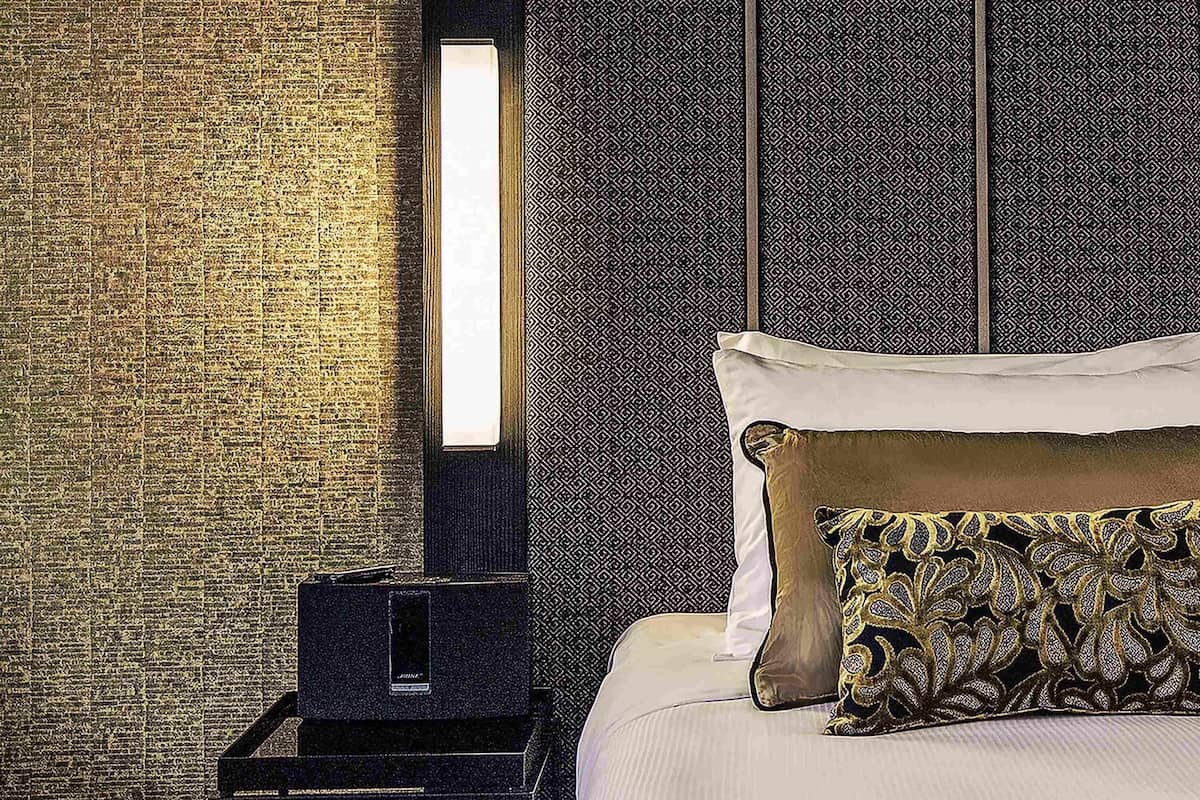 Hotellihuone, jossa on tiilikuviollinen seinä, yöpöytä lampun kanssa ja kuvioiduilla tyynyillä varustettu sänky.