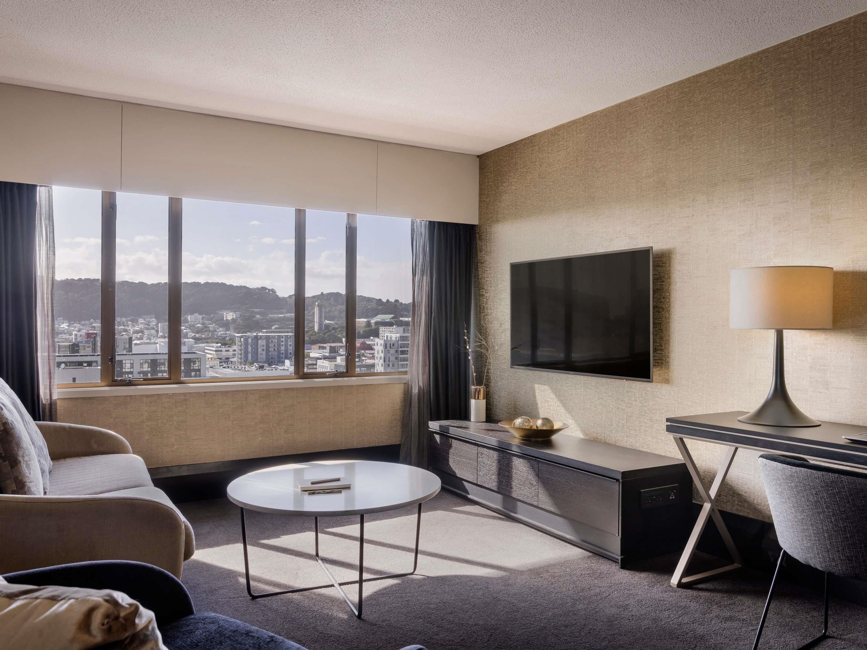Une chambre d’hôtel dotée d’une grande fenêtre, d’un téléviseur à écran plat, d’un bureau, d’une chaise et d’une petite table ronde.