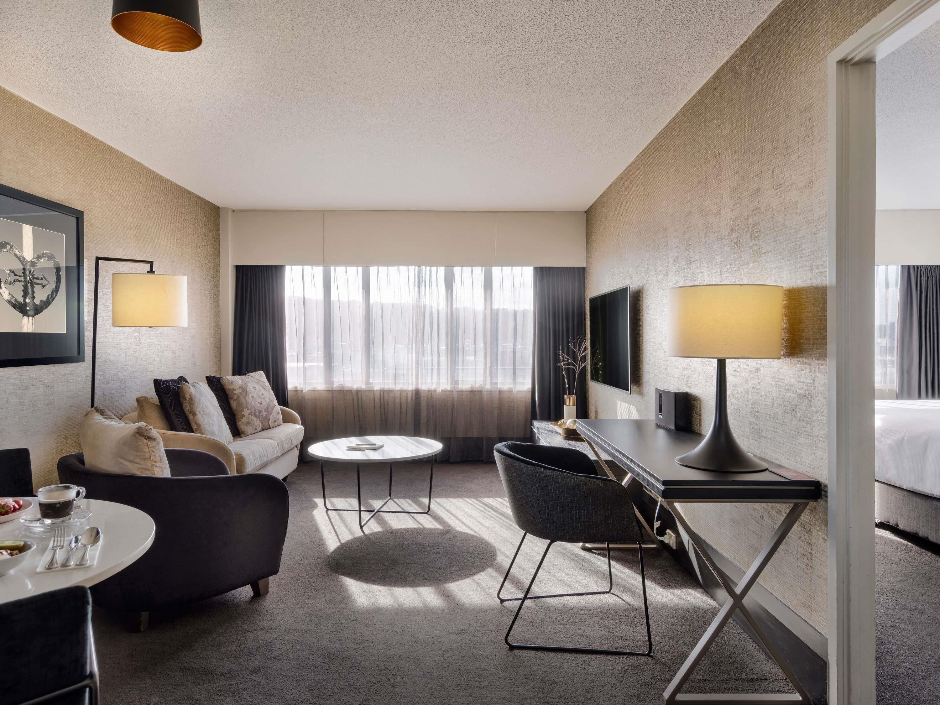 A hotel room with a sofa, a small round table, a desk with a chair, a TV, and a bed.