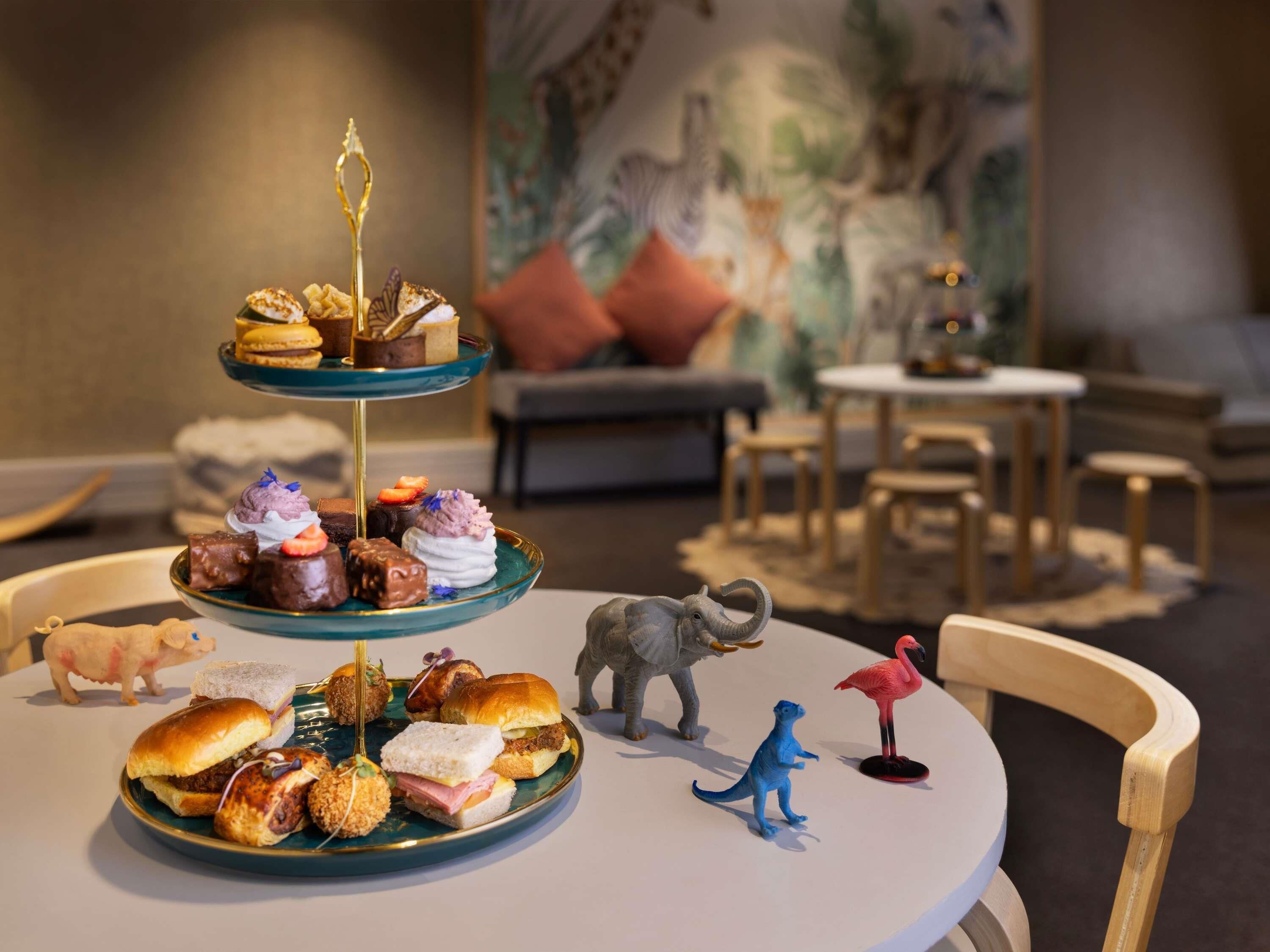 Une table à manger dressée avec une variété de desserts et de sandwichs, entourée de petites figurines de dinosaures et de flamants roses.