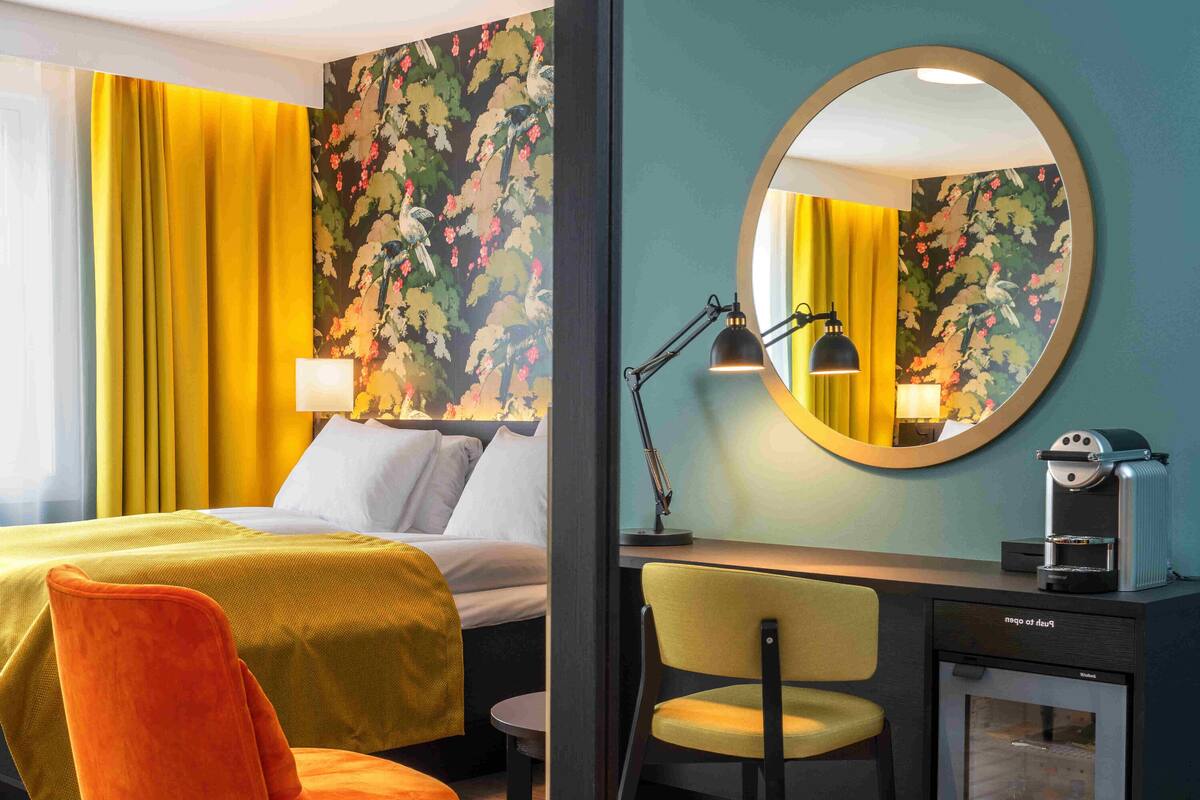 A hotel room with a bed, a desk, a chair, a mirror, and a minibar.