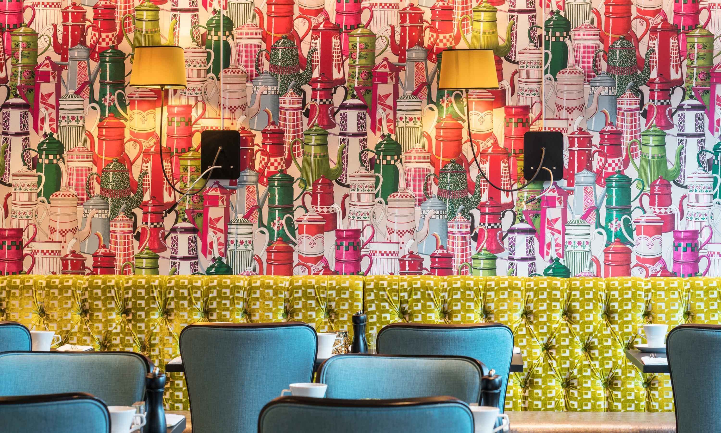 Un restaurant dont un mur est décoré d’une collection de théières colorées et où les tables sont recouvertes d’une nappe jaune à motifs.