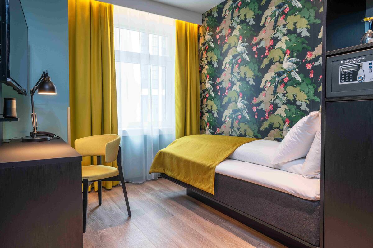 A compact hotel room with a bed, a desk with a lamp, a chair, and a window with yellow curtains.