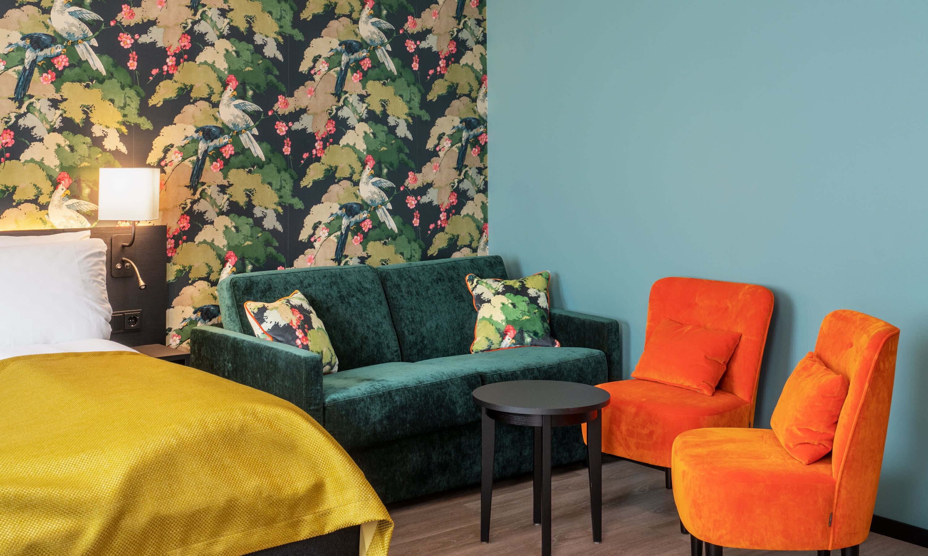 A hotel room with a green sofa, an orange armchair, a small round table, and a bed with a yellow blanket.