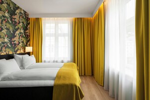 Una habitación de hotel con una cama, un ventanal con cortinas amarillas y un papel tapiz con motivos florales.