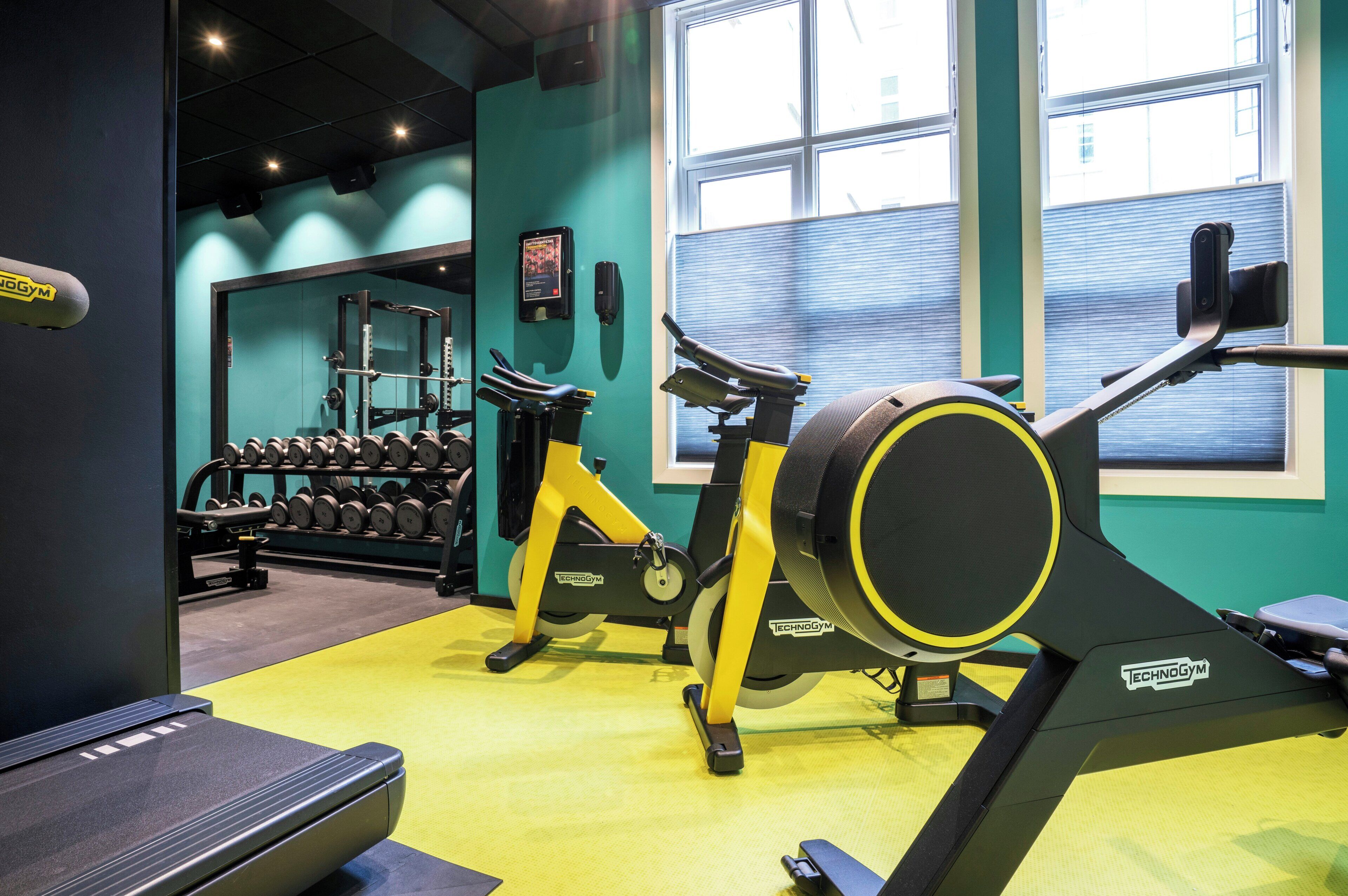 A gym with exercise equipment, a wall-mounted dumbbell rack, and a window with blinds.