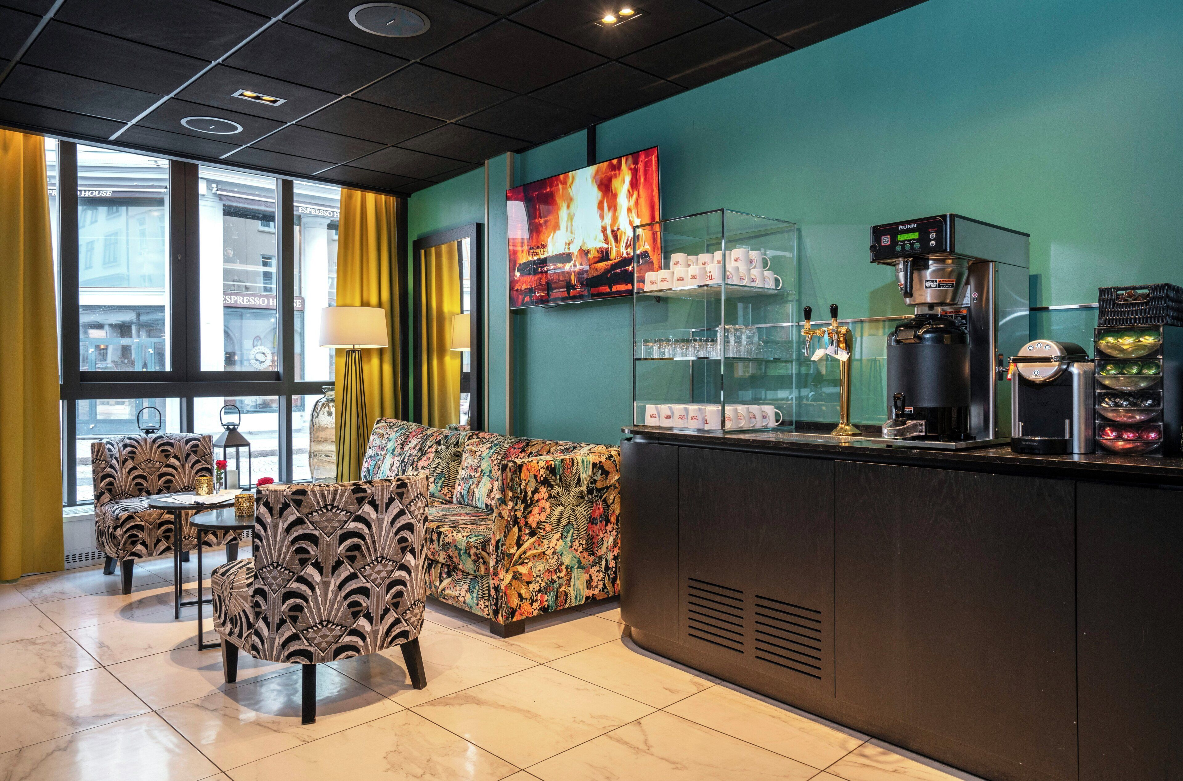 A hotel lobby with a sofa, chairs, a coffee station, and a large window with a view of buildings.