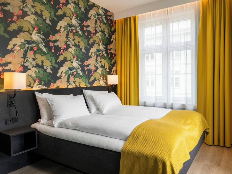 A hotel room with a bed, two bedside lamps, a nightstand, and a window with curtains.