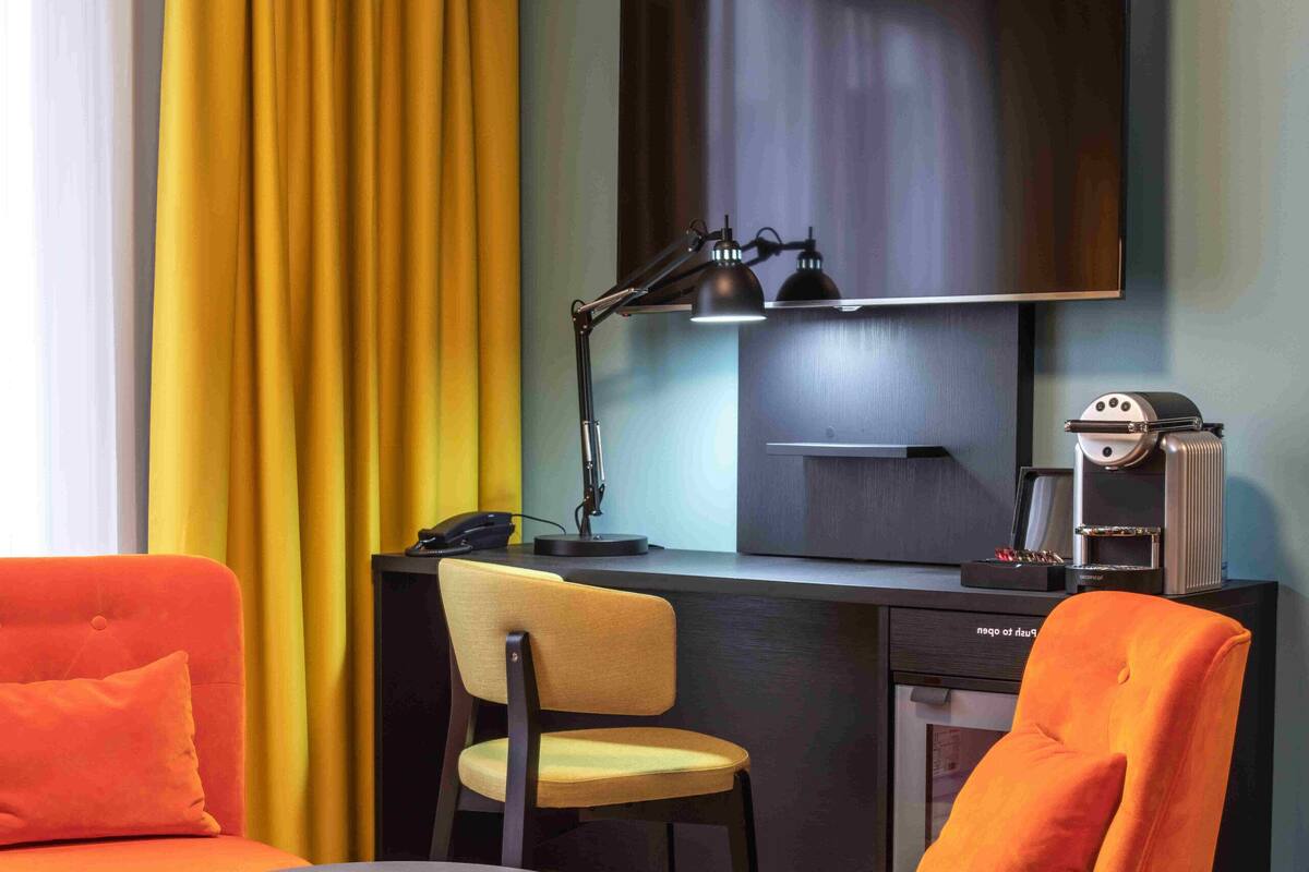 A modern hotel room with a desk, a flat-screen TV, a coffee machine, and vibrant orange and teal furniture.