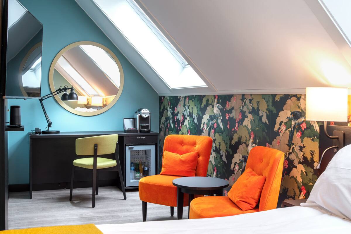 A hotel room with a skylight, a desk with a chair, a bed, and a round mirror.