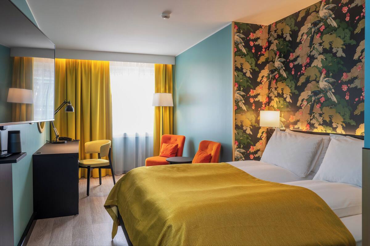 A hotel room with a bed, two chairs, a desk with a lamp, a small table, and a wall with floral wallpaper.