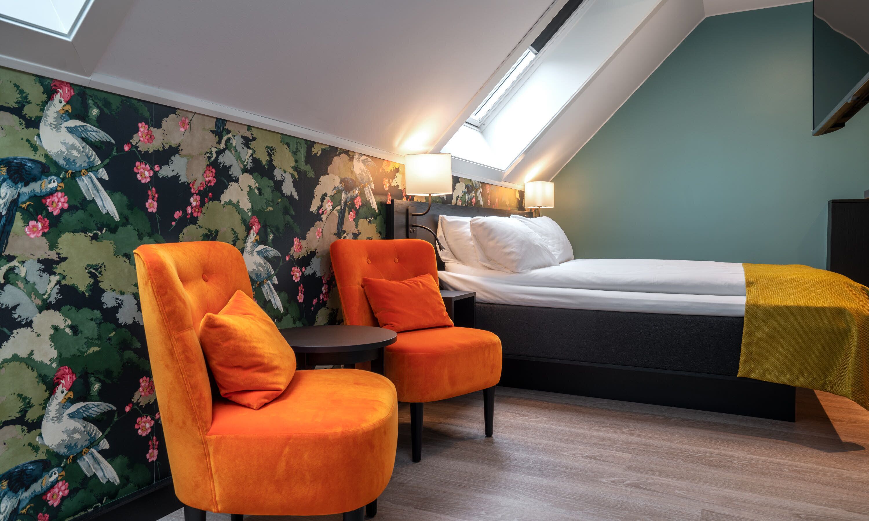 A room with a bed, two orange armchairs, a small table, and a floral wallpaper.