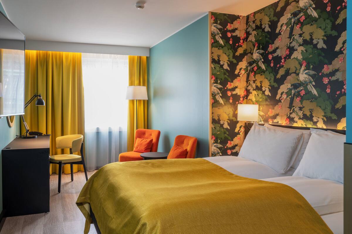 A hotel room with a bed, two chairs, a desk, a lamp, and a television.
