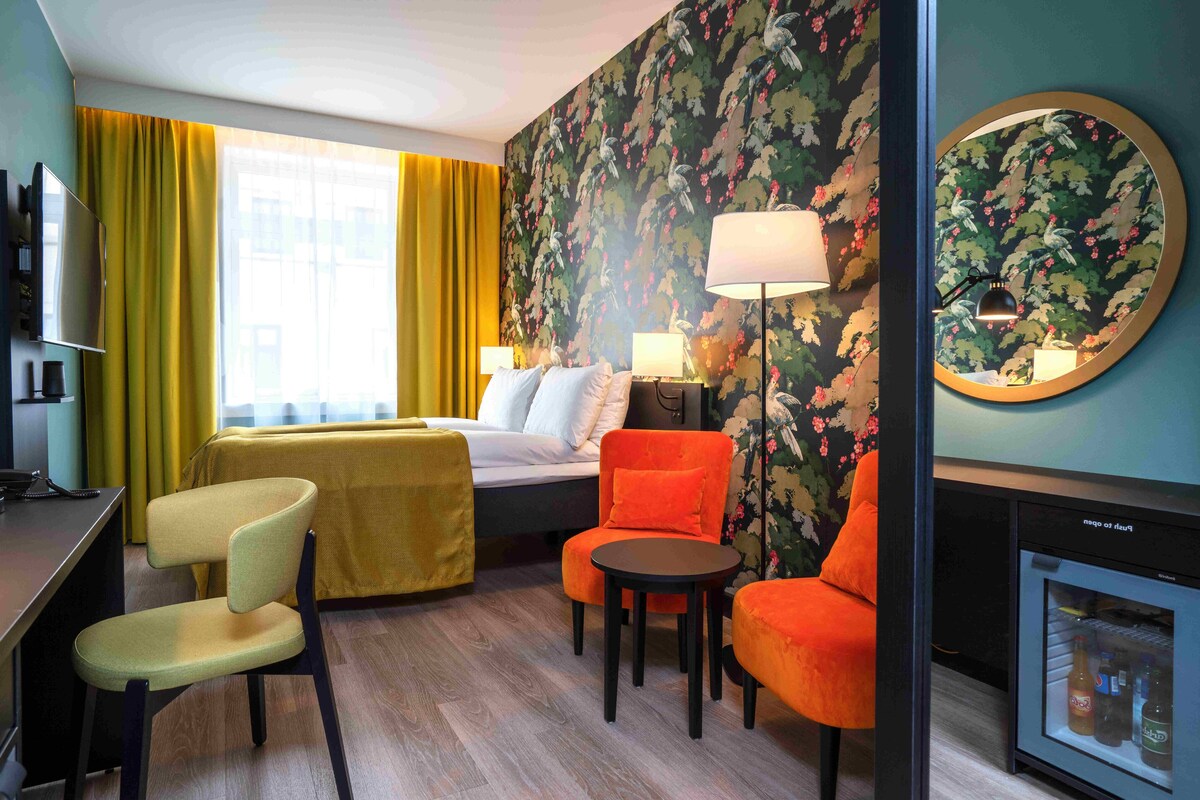 A hotel room with a bed, two chairs, a desk, a TV, a lamp, and a minibar.