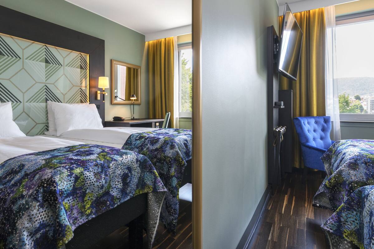 A hotel room with two beds, a TV, a chair, and a window with curtains.