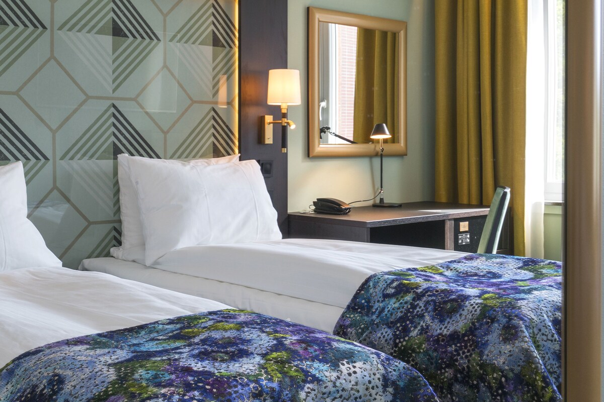 A hotel room with two beds, a desk, a lamp, and a mirror.