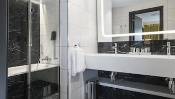 Un baño moderno con ducha envuelta en vidrio, un tocador blanco con salpicadero de mármol negro y un espejo grande con iluminación integrada.