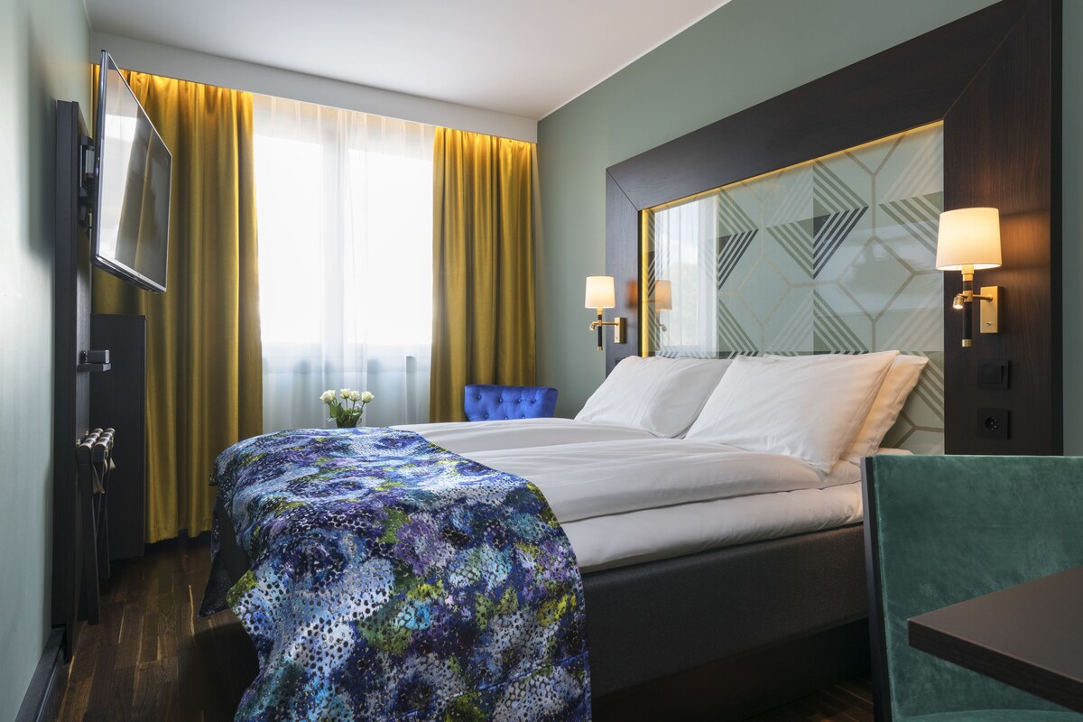 A hotel room with a bed, a desk, a chair, a TV, and a window with curtains.