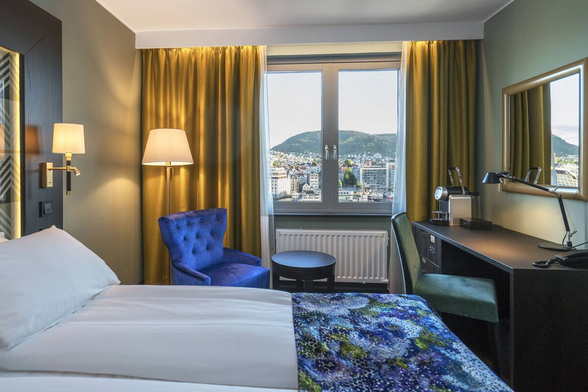 A hotel room with a bed, a blue chair, a desk with a lamp, and a view of a cityscape through a window.