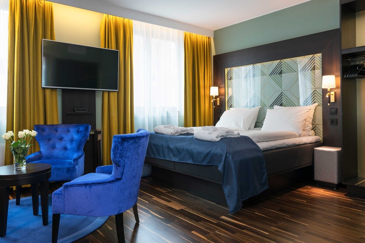 A hotel room with a bed, a TV, a blue armchair, a small table with a vase of flowers, and a bedside lamp.