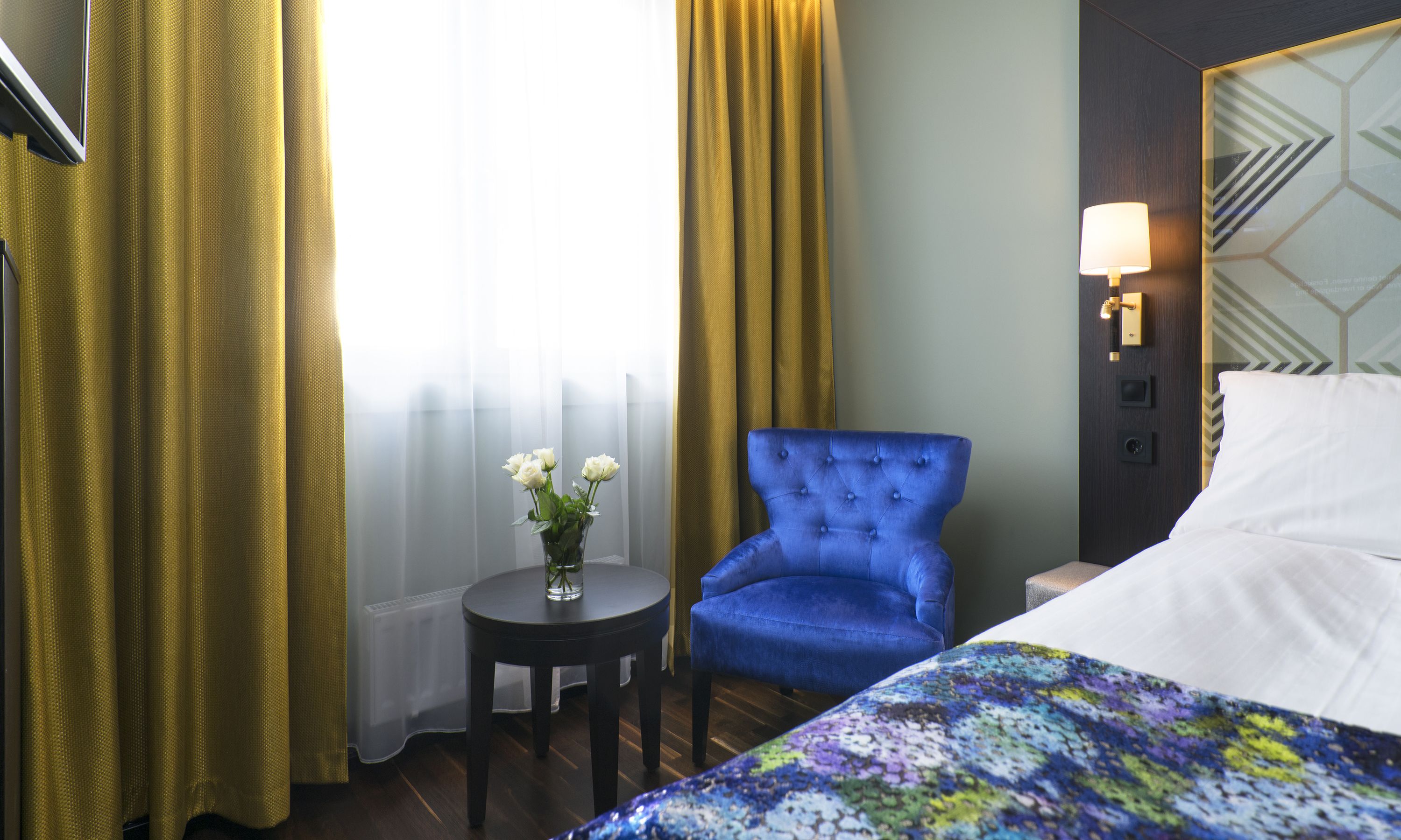 A hotel room with a bed, a blue armchair, a small table with a vase of flowers, and a lamp on the wall.