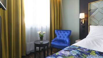 A hotel room with a bed, a blue armchair, a small table with a vase of flowers, and a lamp on the wall.