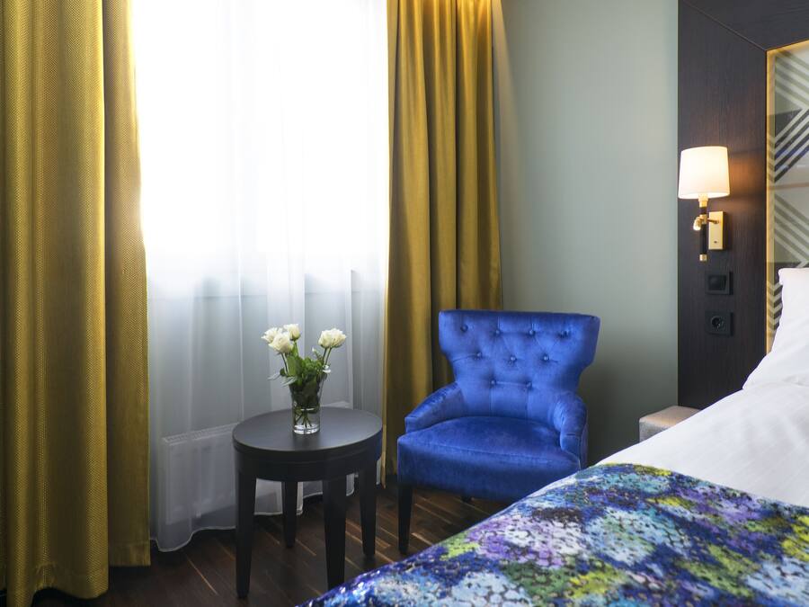 A hotel room with a bed, a blue armchair, a small table with a vase of flowers, and a lamp on the wall.