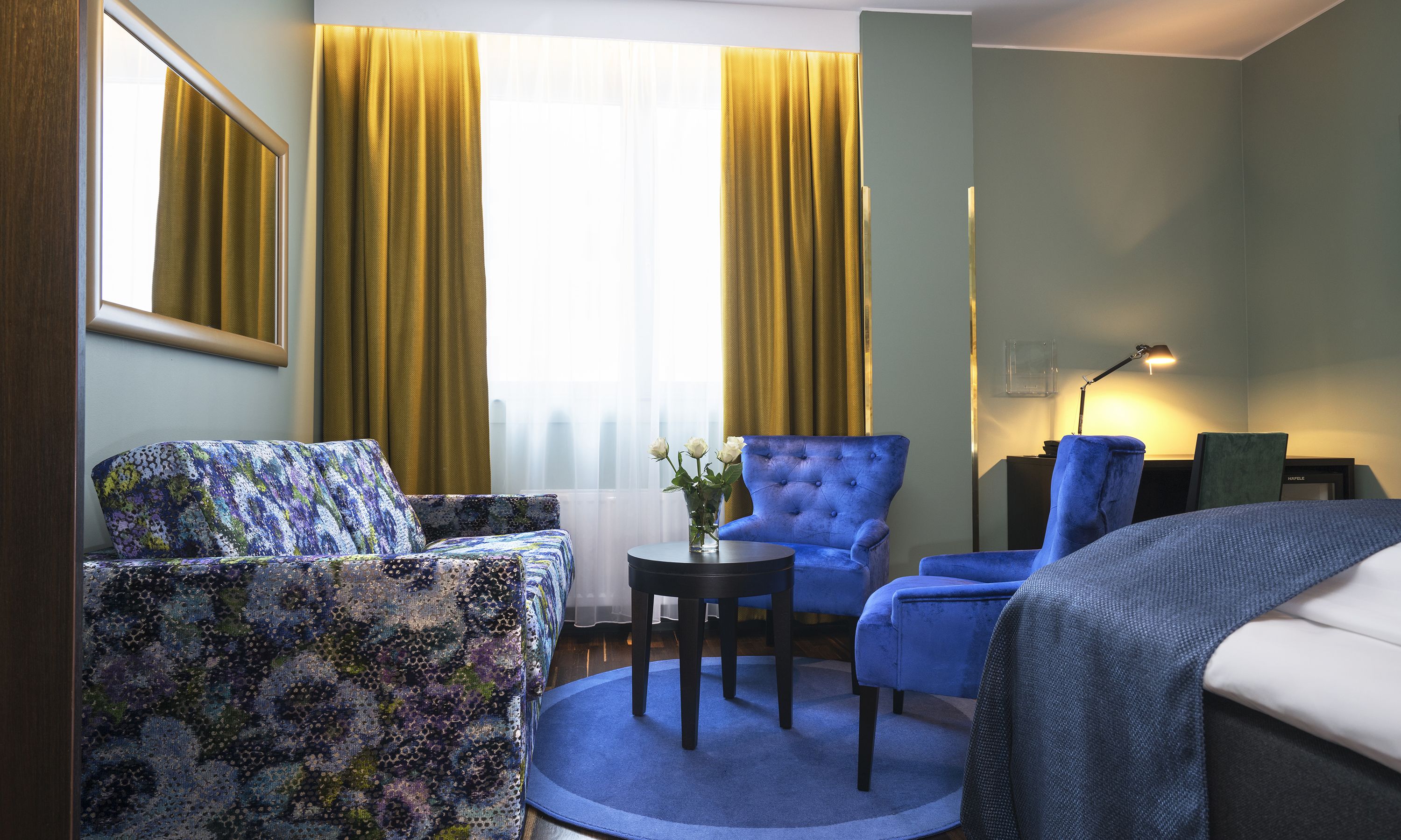 A hotel room with a sofa, two armchairs, a small table, a lamp, and a window with curtains.