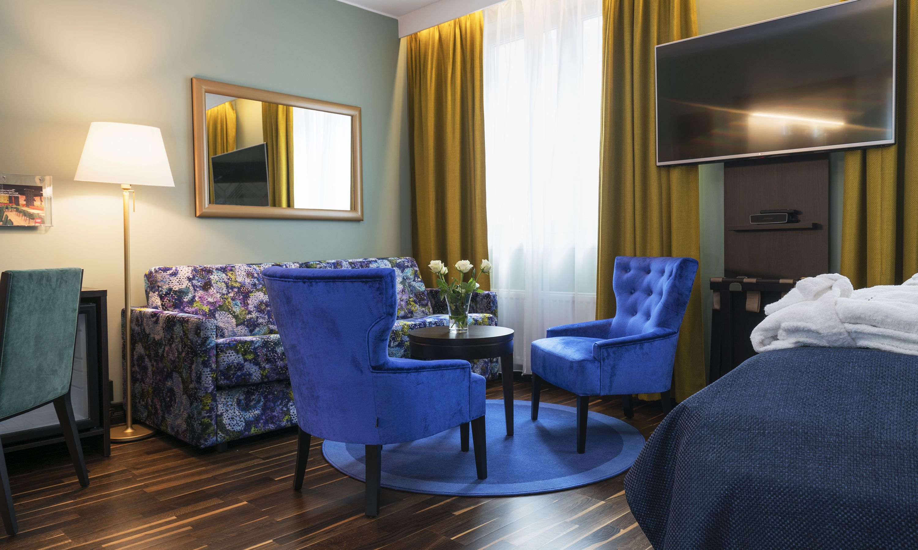Hotelkamer met een bed, twee blauwe fauteuils, een bloemenbank, een klein tafeltje met een bloevas, een flatscreen-tv, een lamp en een raam met gele gordijnen.