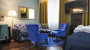 Hotelkamer met een bed, twee blauwe fauteuils, een bloemenbank, een klein tafeltje met een bloevas, een flatscreen-tv, een lamp en een raam met gele gordijnen.