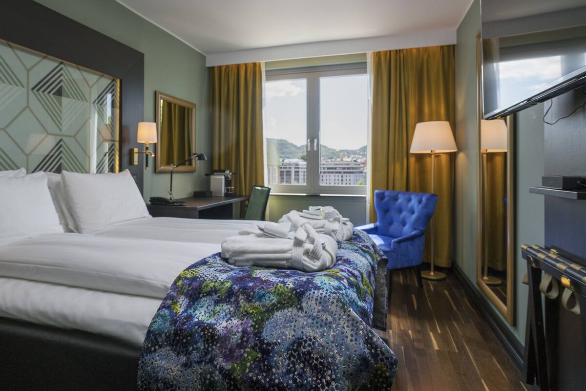 A hotel room with two beds, a blue armchair, a desk with a lamp, and a view of the city through the window.