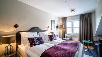 Habitación de hotel con una cama grande, una mesita de noche con lámpara, una pequeña mesa redonda con una taza y una ventana con cortinas.