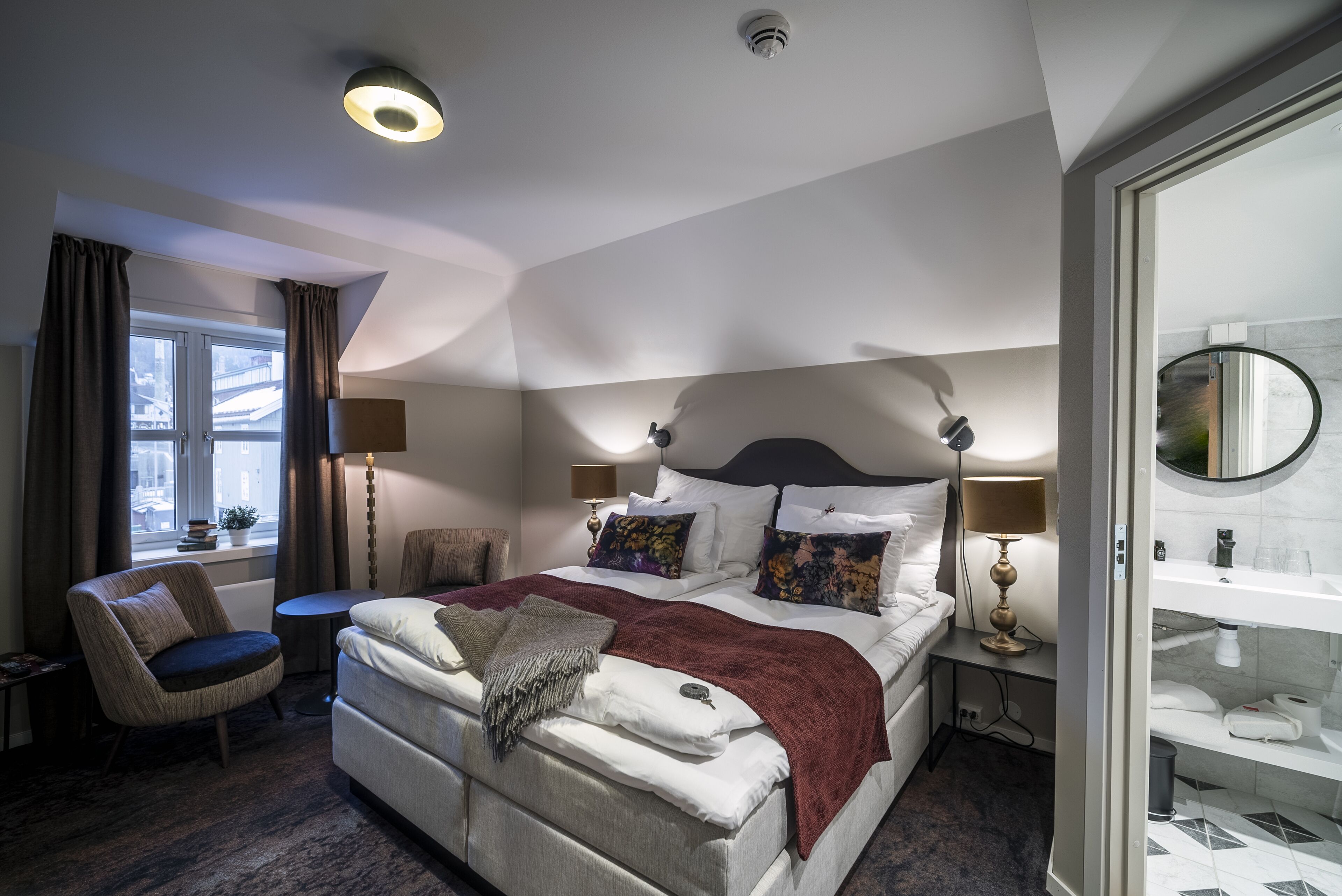 Ein modernes Hotelzimmer mit einem großen Bett, Nachttischen, einem Sessel und einem Badezimmer mit Spiegel und Ablageflächen.