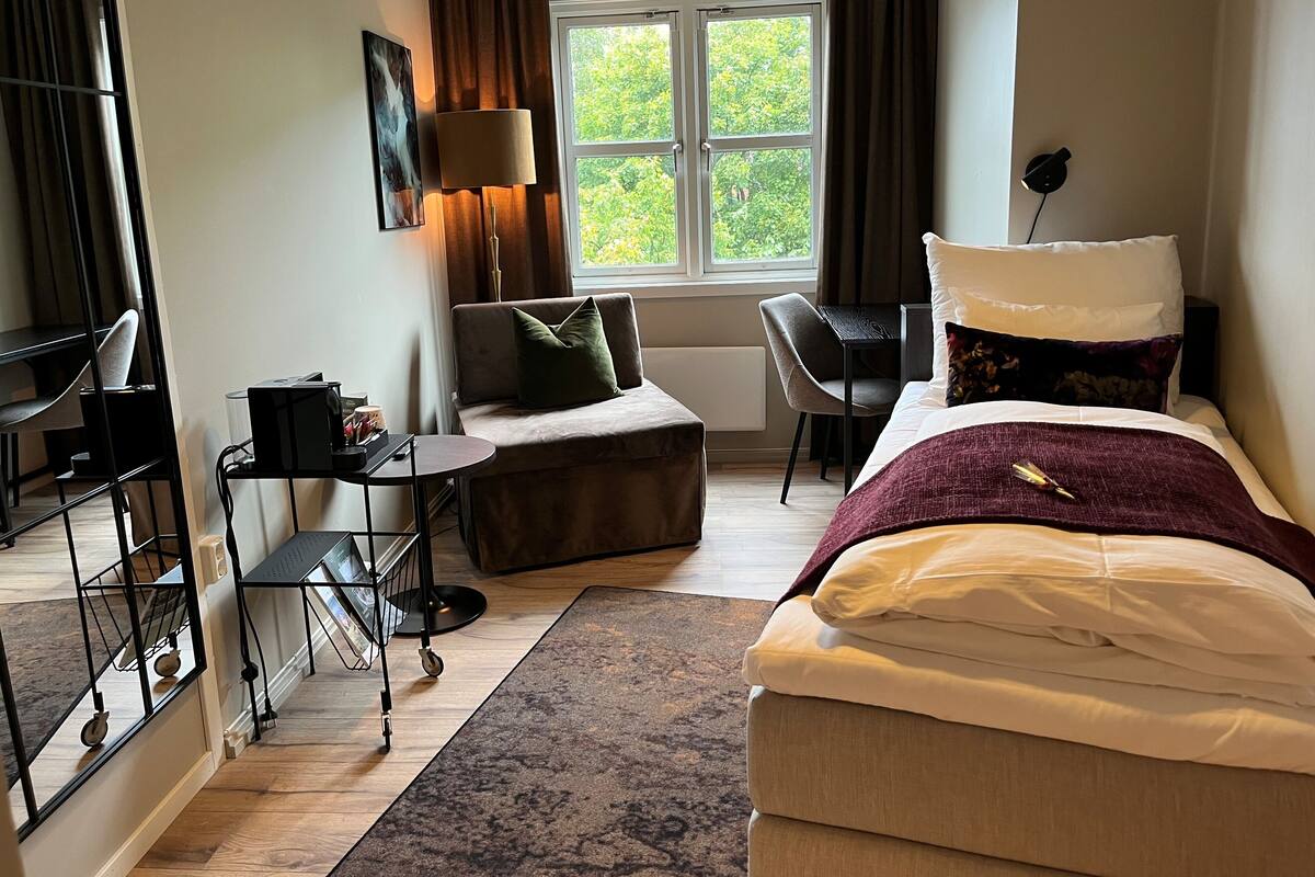 A hotel room with a bed, a desk, a chair, a sofa, and a window with a view of trees.
