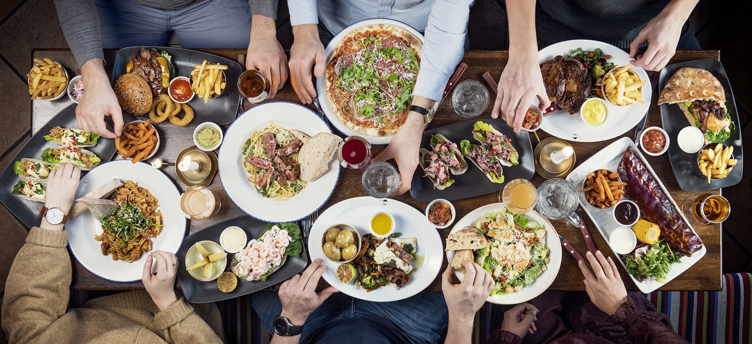 A group of people gathered around a table with various dishes, including pasta, burgers, and salads.