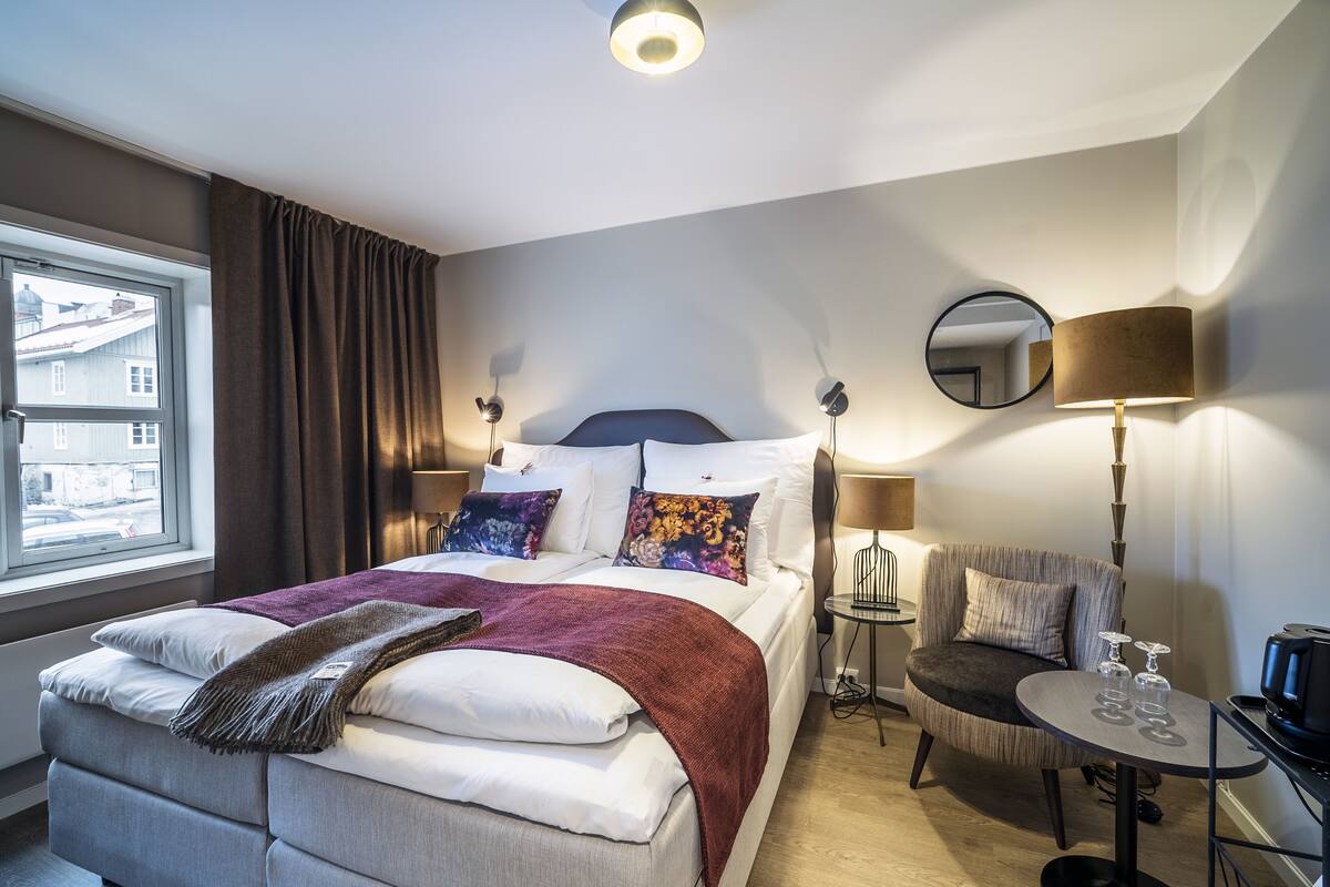 A hotel room with a bed, bedside tables, a chair, a round table, and a window.