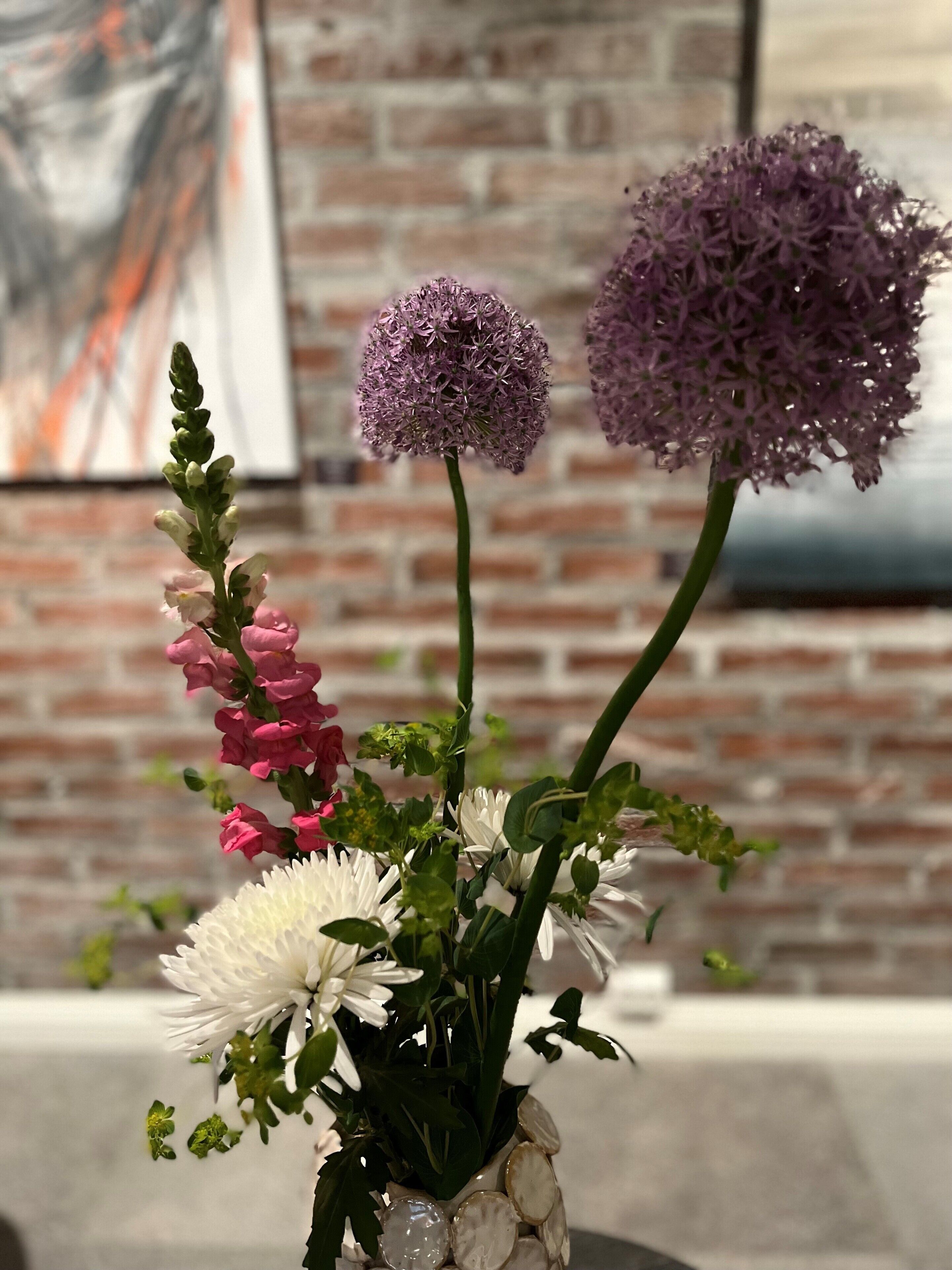 Un ramo de flores en un jarrón con un muro de ladrillo y un cuadro al fondo.