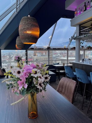 A modern bar area with a city view, a large vase of flowers, and hanging pendant lights. - Aksjemøllen - by Classic Norway Hotels (Lillehammer)