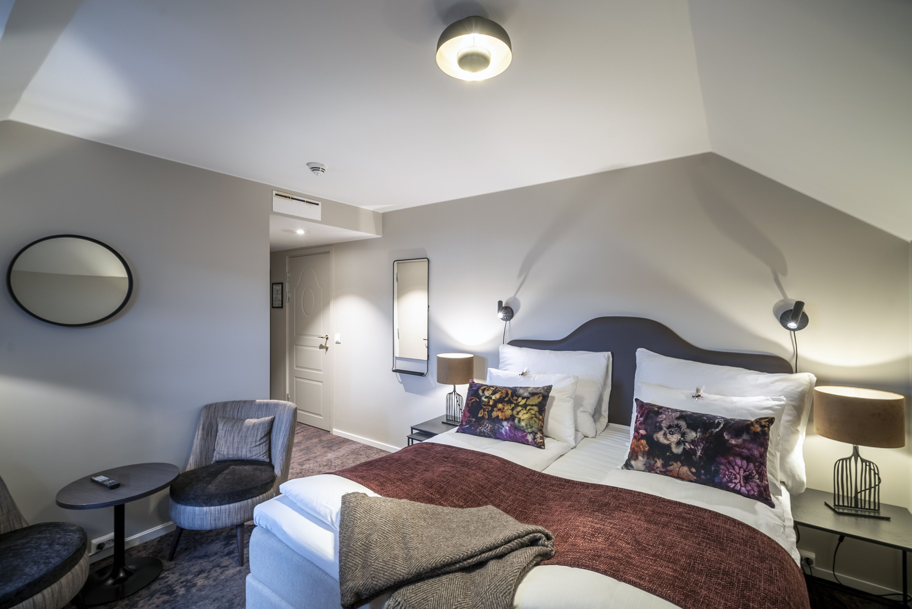 A hotel room with a large bed, a round table, a chair, a desk, and a mirror.